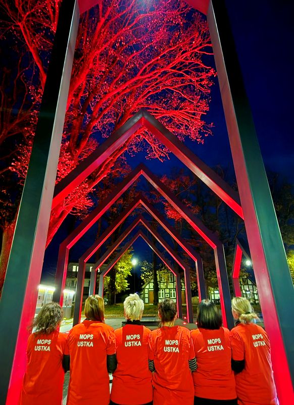 „Grupa sześciu osób stoi tyłem, obejmując się ramionami. Wszyscy mają na sobie czerwone koszulki z napisem ‘MOPS USTKA’. Znajdują się na tle iluminowanej na czerwono instalacji artystycznej i drzew, oświetlonych nocą w intensywnych czerwonych barwach.”