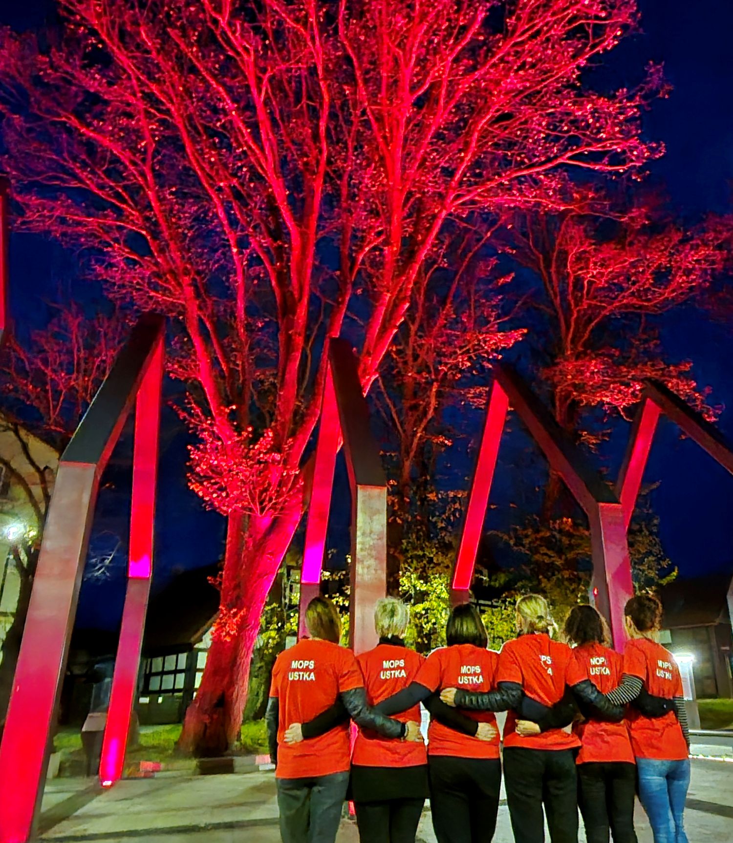 „Grupa sześciu osób stoi tyłem, obejmując się ramionami. Wszyscy mają na sobie czerwone koszulki z napisem ‘MOPS USTKA’. Znajdują się na tle iluminowanej na czerwono instalacji artystycznej i drzew, oświetlonych nocą w intensywnych czerwonych barwach.”