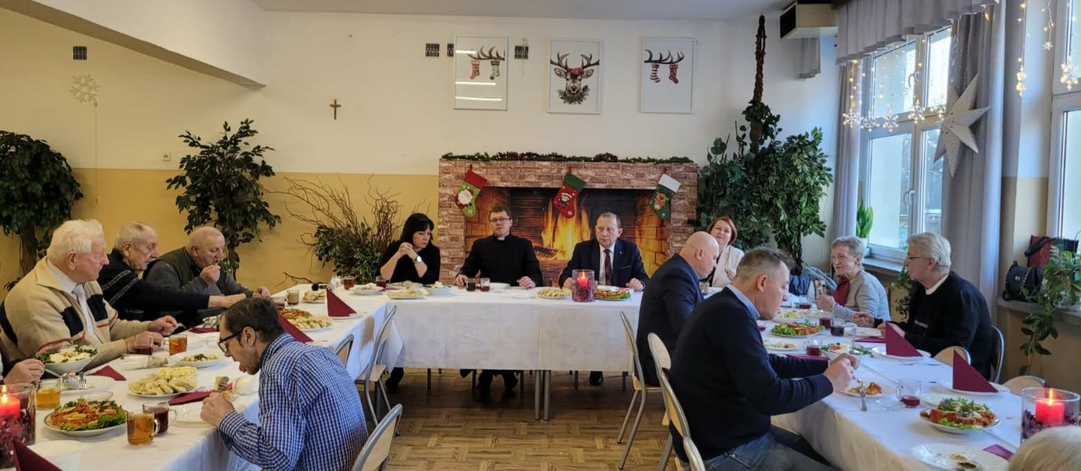 Grupa starszych i młodszych osób siedzi przy długich stołach, wspólnie jedząc świąteczny posiłek w jasnej sali. W tle widać dekoracje bożonarodzeniowe: kominek z girlandą i skarpetami, lampki, choinkowe ozdoby oraz obrazki z motywem reniferów. Na stołach stoją talerze z jedzeniem, napoje i świece, a atmosfera jest spokojna i uroczysta.