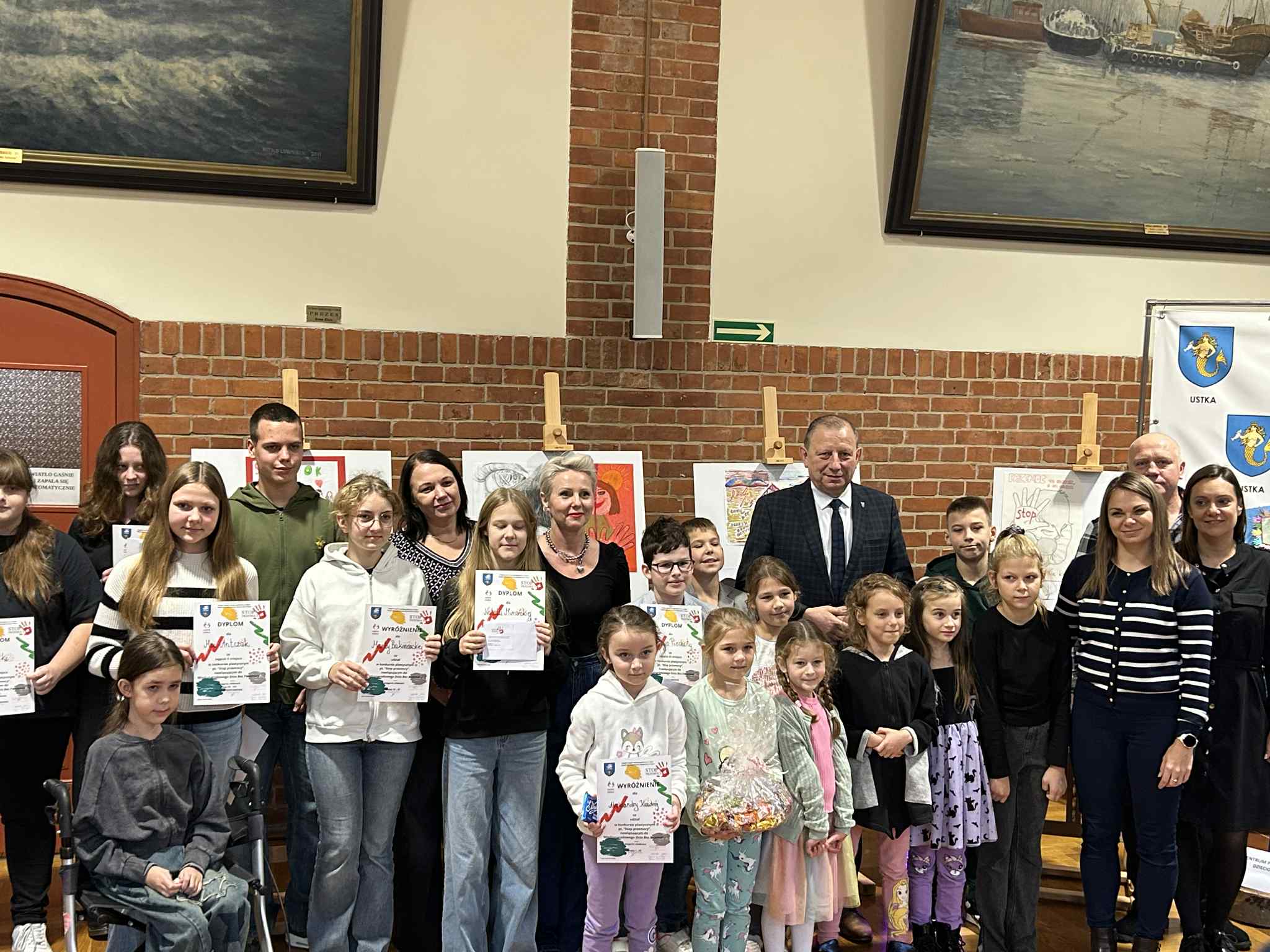 Grupa dzieci, młodzieży i dorosłych pozuje do wspólnego zdjęcia w sali z ceglanymi ścianami. Część osób trzyma dyplomy, a w tle widać prace plastyczne ustawione na sztalugach. Po prawej stronie stoją przedstawiciele organizatorów wydarzenia, a w tle widoczne są obrazy marynistyczne oraz herb miasta Ustka