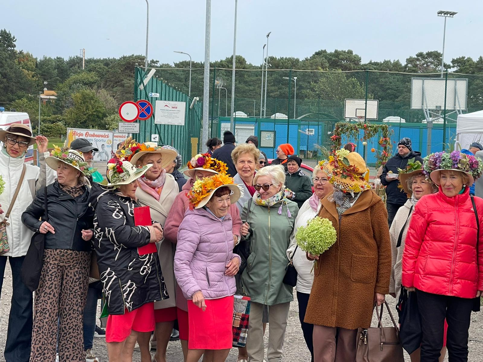 Grupa starszych kobiet ubranych w kolorowe stroje i fantazyjne kapelusze ozdobione kwiatami oraz liśćmi pozuje do zdjęcia na świeżym powietrzu podczas wydarzenia plenerowego. W tle widać boisko sportowe, namioty oraz innych uczestników. Atmosfera jest radosna i festiwalowa.