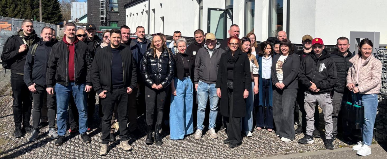 rupa kilkunastu osób stoi na zewnątrz przed nowoczesnym budynkiem, ustawiona do wspólnego zdjęcia. Uczestnicy są ubrani głównie w kurtki i ubrania w ciemnych kolorach, co sugeruje chłodniejszą pogodę. W tle widać fragmenty zabudowy miejskiej i ogrodzenie. Większość osób patrzy w stronę aparatu, niektórzy się uśmiechają.