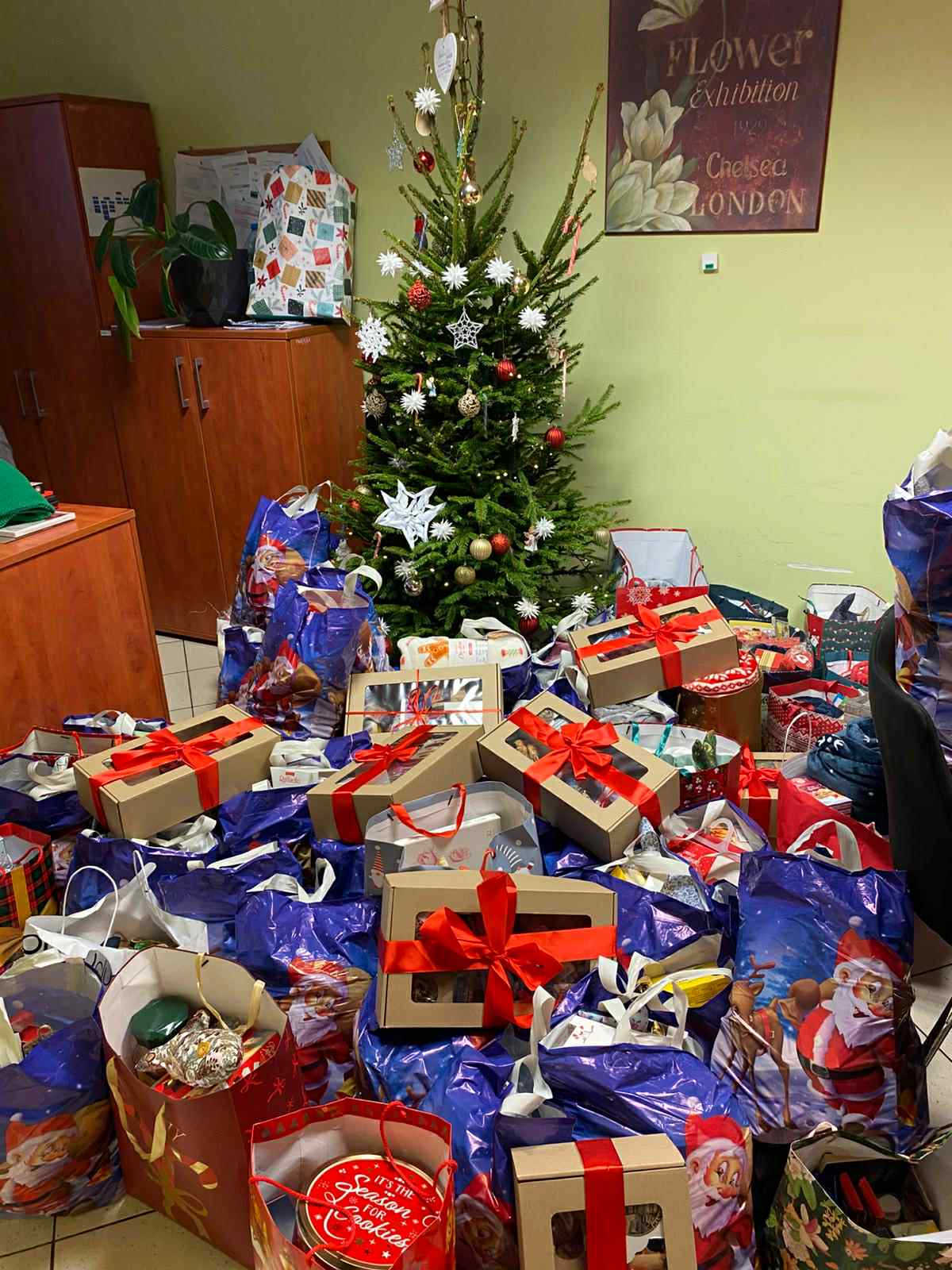 „Udekorowana choinka stojąca w pomieszczeniu, otoczona dużą liczbą świątecznych prezentów i toreb z podarunkami; wokół choinki leżą pudełka z czerwonymi wstążkami i torby z motywami bożonarodzeniowymi